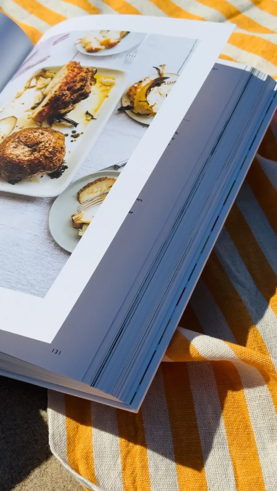Opengeslagen kookboek met een foto van geroosterde groenten, liggend op een geel-wit gestreepte handdoek in de zon